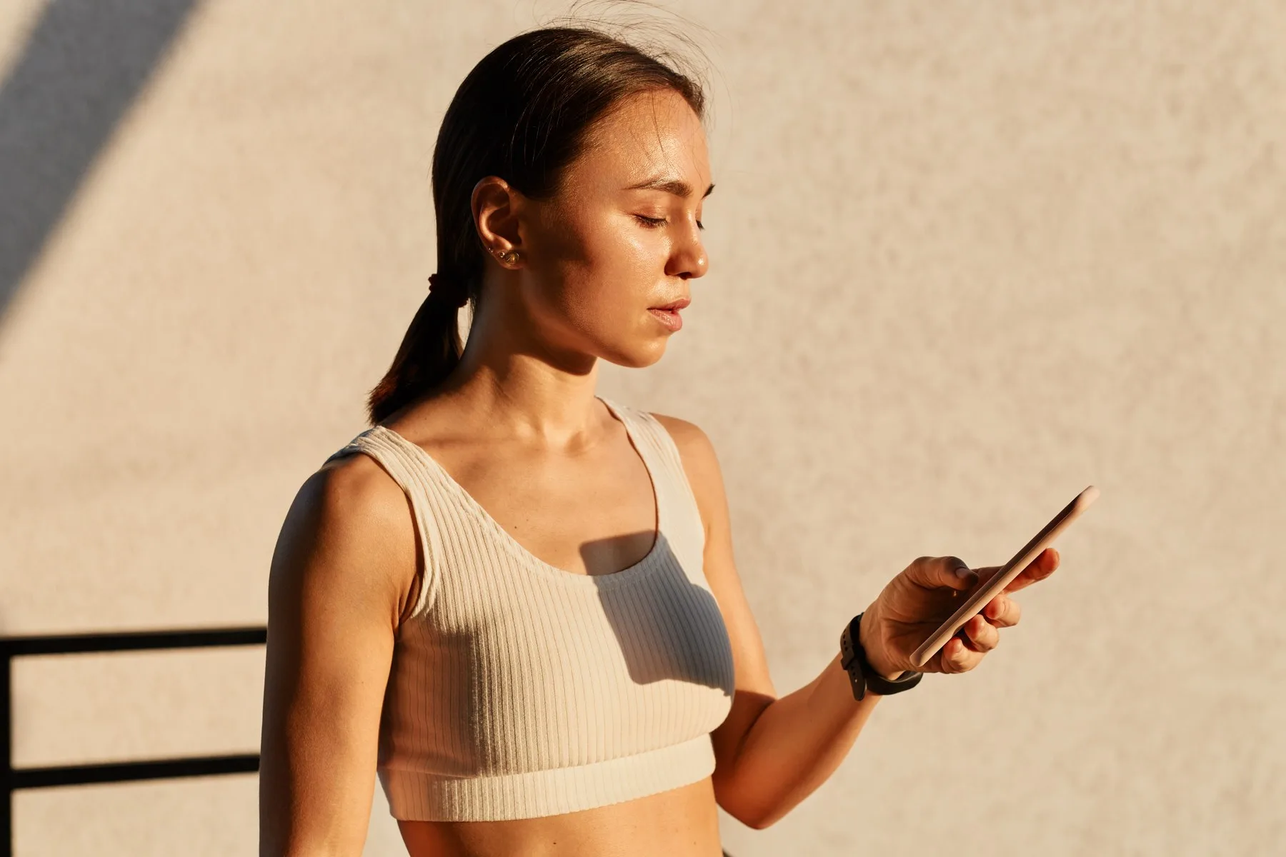 Une femme en brassière sportive claire consulte son téléphone au soleil couchant, devant un mur clair